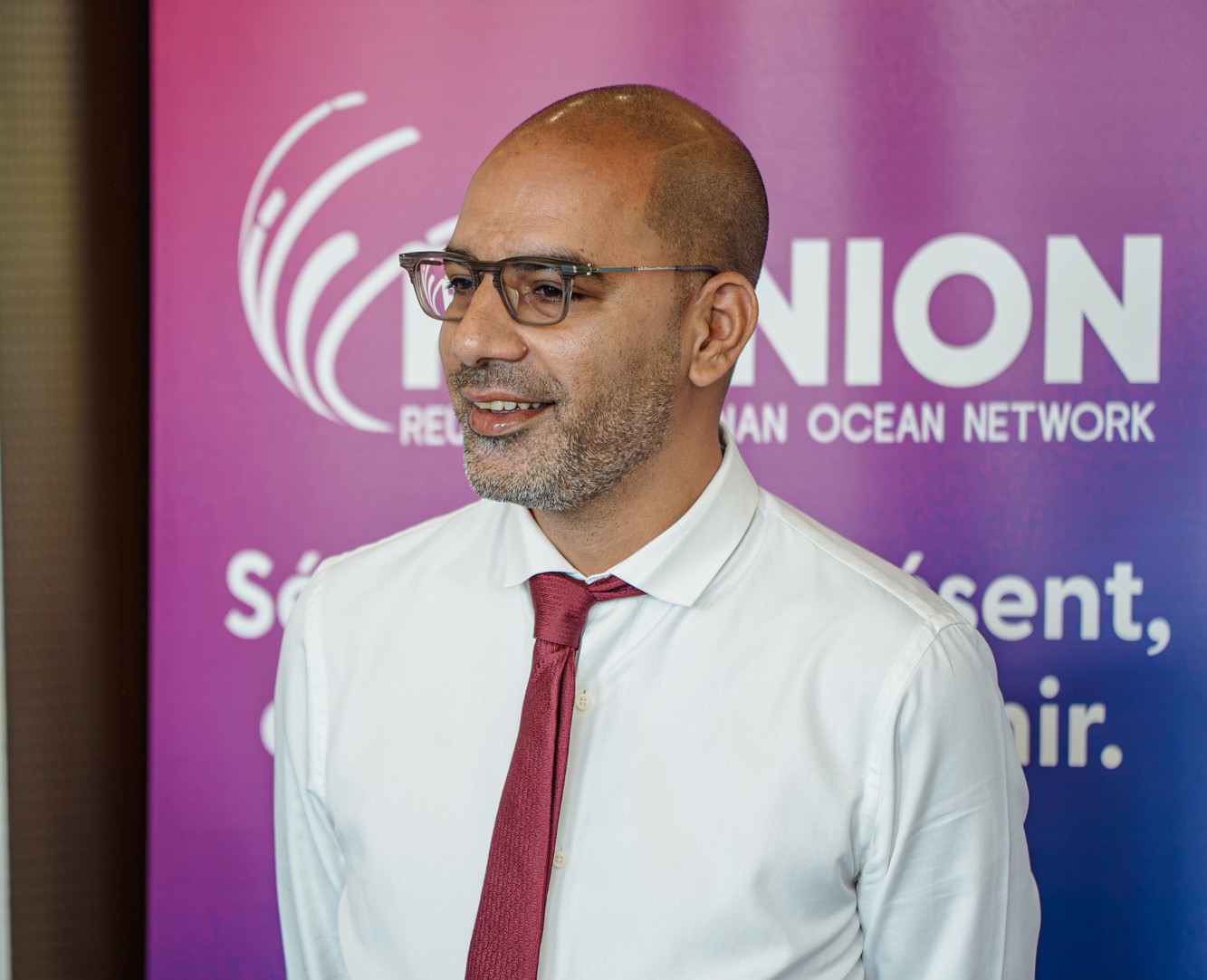 Un homme portant une chemise blanche et une cravate bordeaux devant une affiche promotionnelle Réunion Ocean Network.